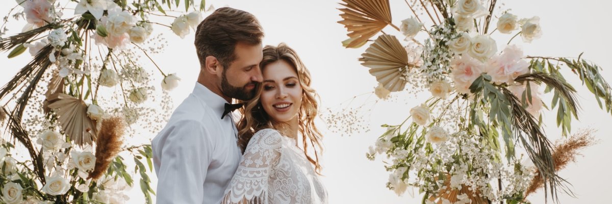 Ein Mann in einem Anzug hält eine Frau in einem weißen Kleid in den Armen vor einem großen Blumenbogen 