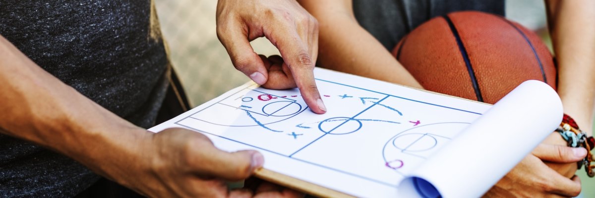 Einer hält ein Klemmbrett in der Hand wo Spielanweisungen drauf gezeichnet sind, ein anderer hält einen Basketball in den Händen