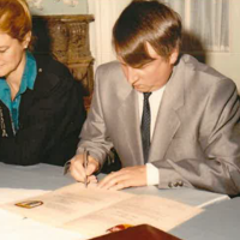 Ein Mann und eine Frau sitzen an einem Tisch und und vor dem Mann liegen Blätter und er hat einen Stift in der Hand 