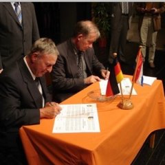 Zwei Männer sitzen an einem Tisch und schreiben etwas auf ein Blatt. Vor Ihnen steht eine kleine Deutschland Flagge und eine Niederländische Flagge