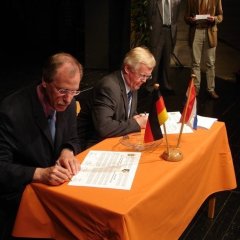 Zwei Männer sitzen an einem Tisch und schreiben etwas auf ein Blatt. Vor Ihnen steht eine kleine Deutschland Flagge und eine Niederländische Flagge