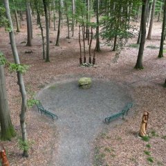 Man sieht einen Platz im Wald an dem einige Bänke stehen und in der Mitte steht ein großer Stein und dahinter ist ein großes Kreuz an den Bäumen befestigt. Am Platzeingang stehen jeweils rechts und links eine Figur aus Holz (Ansicht von oben) 