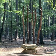 Kreuz hängt an vier Baumstämmen und davor liegt ein großer Stein