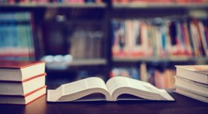 Ein aufgeschlagenes Buch liegt auf dem Tisch und im Hintergrund ist ein großes Bücherregal