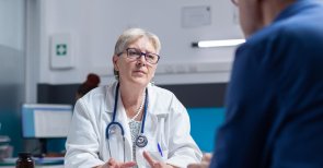 Eine Frau mit einem weißen Kittel und einem Stethoskop um den Hals hat unterlagen vor sich liegen und vor ihr sitzt ein Mann