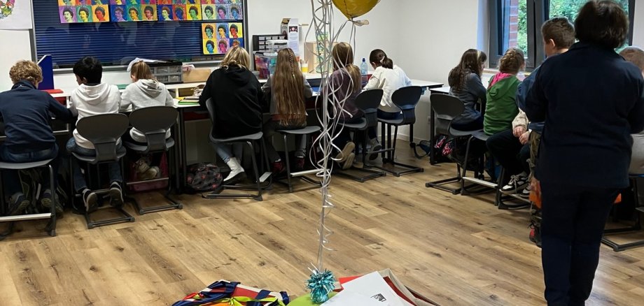 Im Vordergrund stehen Kisten in einem Kreis mit Ballons in der Mitte und im Hintergrund sitzen einige Kinder an einem Tisch 