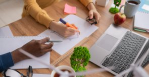 Ein Schreibtisch auf dem ein Laptop steht und einige Zettel liegen und an dem zwei Menschen sitzen