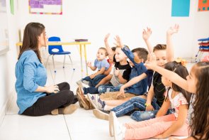 Frau sitzt vor einigen Kleinen Kindern auf dem Boden und die Kinder heben die Hand